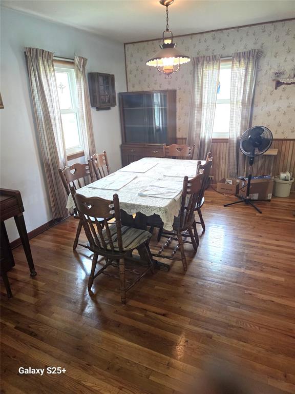 202 E Main St 04 A dining room with a wooden table covered by a white lace tablecloth, six wooden chairs, hardwood floors, two windows with curtains, a standing fan, boxes, and a cabinet in the corner.