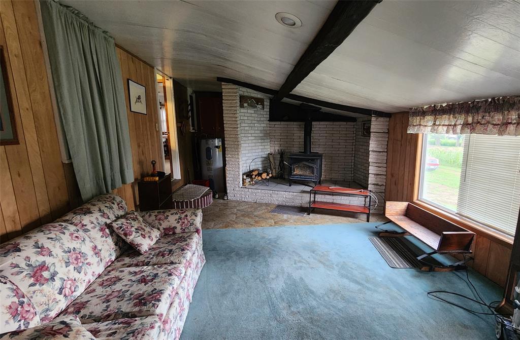 202 E Main St 06 A cozy living room with wood-paneled walls, floral couch, blue carpet, a wood stove set against a white brick wall, large windows with curtains, and a bench below the windows letting in natural light.