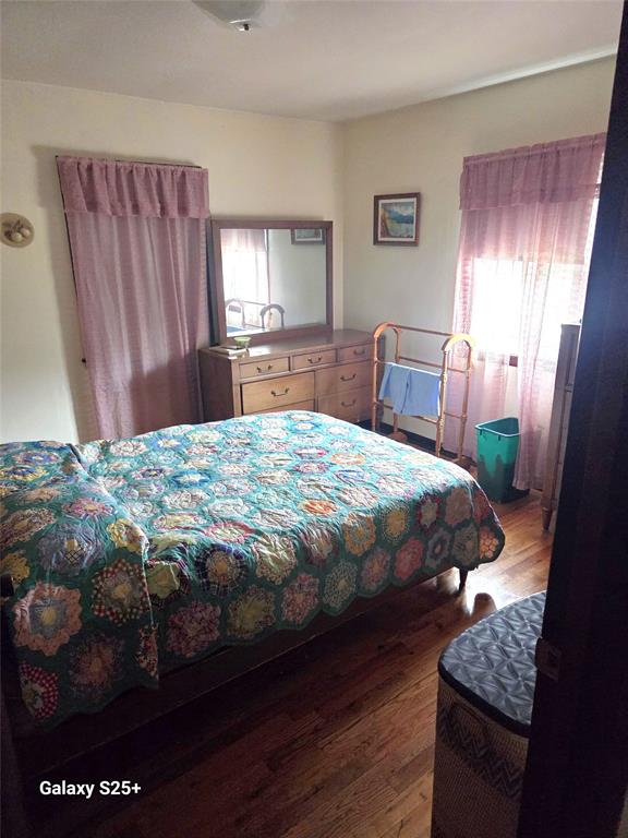 202 E Main St 07 A cozy bedroom with a colorful patterned quilt on the bed, wooden dresser and mirror, walker, laundry basket, green trash bin, and lace curtains on two windows letting in natural light.