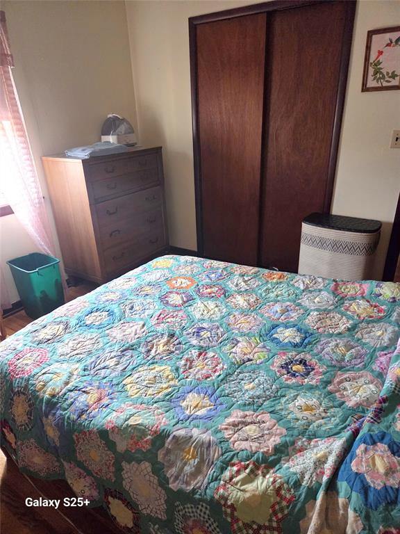 202 E Main St 08 A bedroom with a colorful floral quilt on the bed, a wooden dresser with items on top, a laundry basket, a window with blinds, and a green trash bin in the corner.