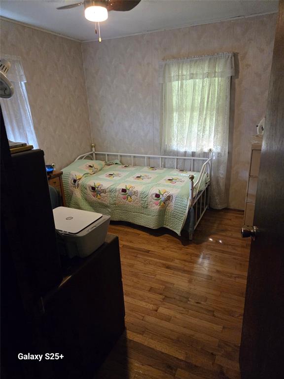 202 E Main St 09 A small bedroom with a single metal bed frame, a light-colored quilt, wood floors, a window with sheer curtains, and a dresser. A ceiling fan is partially visible above, and the door is open.