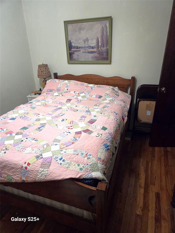 202 E Main St 10 A wooden bed with a colorful, patterned quilt sits in a small room with hardwood floors. A lamp and framed painting are on the wall, and a cardboard box is on a chair next to the bed. "Galaxy S25+" text is visible.