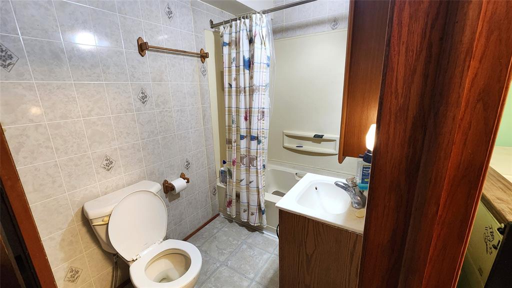 202 E Main St 11 A small bathroom with a toilet, open lid, wall-mounted toilet paper, a shower with a patterned curtain, a sink with a cabinet and mirror, soap dispenser, and a lit bulb above the sink. The walls and floor are tiled.