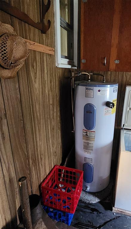 202 E Main St 14 A hot water heater is installed in a small room with wood-paneled walls. A red plastic crate sits on the floor nearby, and a straw hat hangs on a hook next to a window and wooden cabinets.