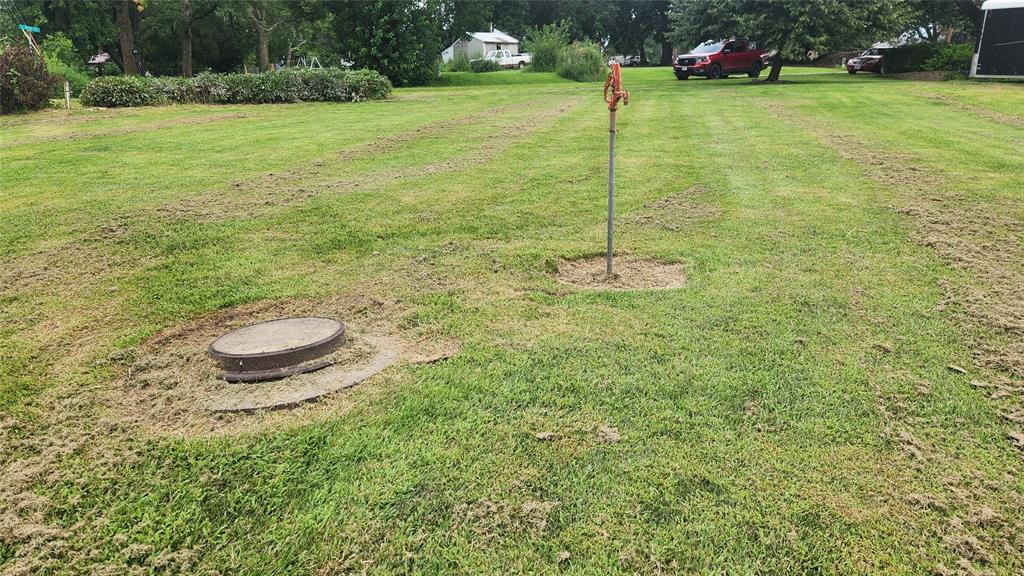 202 E Main St 15 A neatly mowed grassy yard with a round, metal septic cover on the left and a tall metal pole with an orange marker on the right. Some grass is left uncut around both objects. Trees and vehicles are in the background.