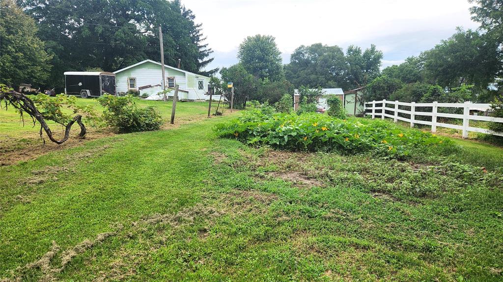 202 E Main St 16 A grassy yard with a white house in the background, a fenced garden with green plants and flowers, a white wooden fence to the right, and trees surrounding the area under a cloudy sky.