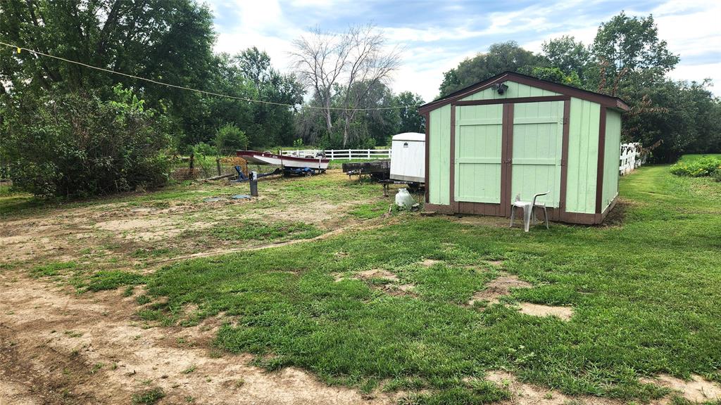 202 E Main St 18 A small green shed stands on grass near a white fence, with a white trailer and a boat in the background. A white plastic chair sits beside the shed, and trees surround the area under a partly cloudy sky.