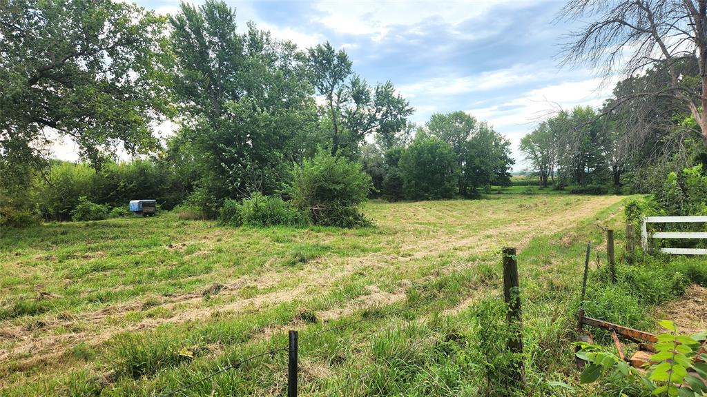 202 E Main St 22 A grassy, partially mowed field bordered by trees under a cloudy sky. A small blue structure sits in the distance on the left, and a white fence and wire fencing are visible in the foreground on the right.