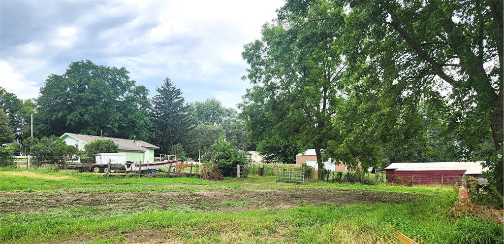 202 E Main St 24 A rural scene with a grassy yard, trees, a small house, farm equipment, fences, and a red barn under a cloudy sky.