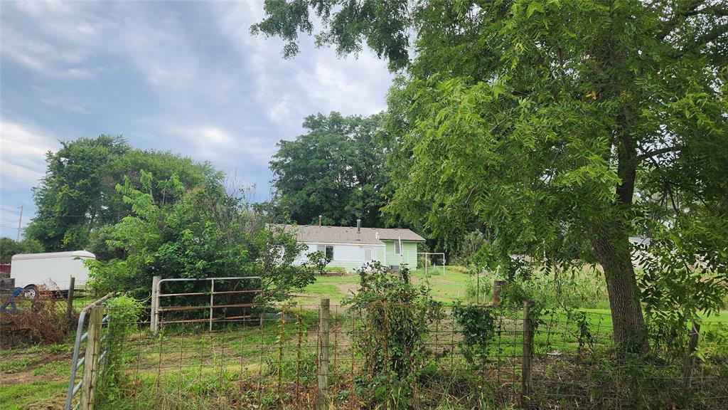 202 E Main St 25 A small white house is partially hidden by trees and bushes in a green, rural yard. A wire fence and metal gate are in the foreground, with a white trailer to the left and a cloudy sky above.