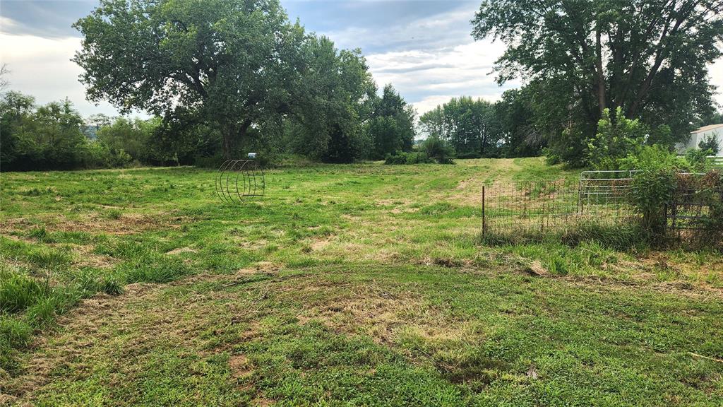 202 E Main St 27 A grassy, open field with patches of uneven grass, surrounded by tall trees under a partly cloudy sky. There are metal animal pens and fencing on the right side of the image.