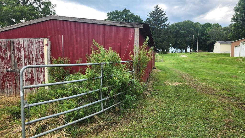 202 E Main St 28 A red wooden shed with a weathered door stands beside a partially open metal gate, surrounded by overgrown greenery. A grassy path leads uphill toward trees and other buildings in the background.