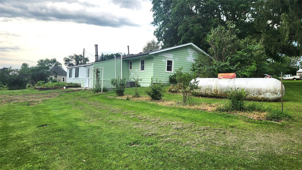 202 E Main St 30 A light green single-story house sits on a grassy lawn with a large white propane tank in the foreground and bushes along the side of the house; trees and a cloudy sky are in the background.