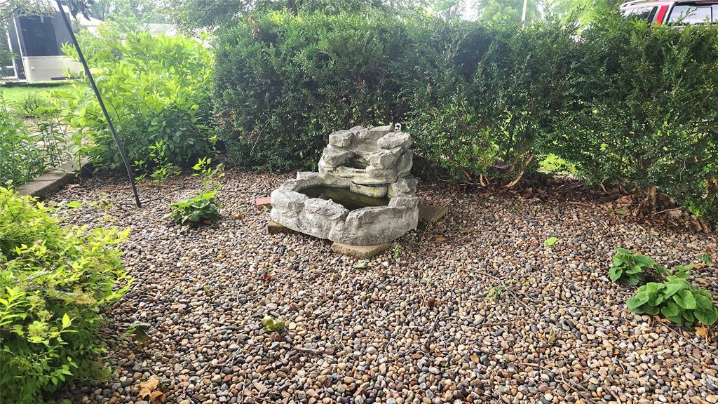 202 E Main St 31 A small stone water fountain sits surrounded by pebbles and green shrubs in a garden area, with leafy plants and bushes in the background.