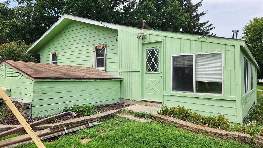 202 E Main St 32 A small, light green house with a slanted roof, diamond-patterned window on the door, large front window, and attached porch. The yard has some overgrown plants and pipes lying on the grass.