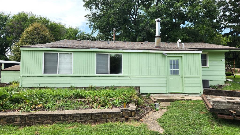 202 E Main St 33 A light green single-story house with a small garden bed in front, three windows, a gray door, and a sloped shingle roof, set against a backdrop of tall green trees and grass.
