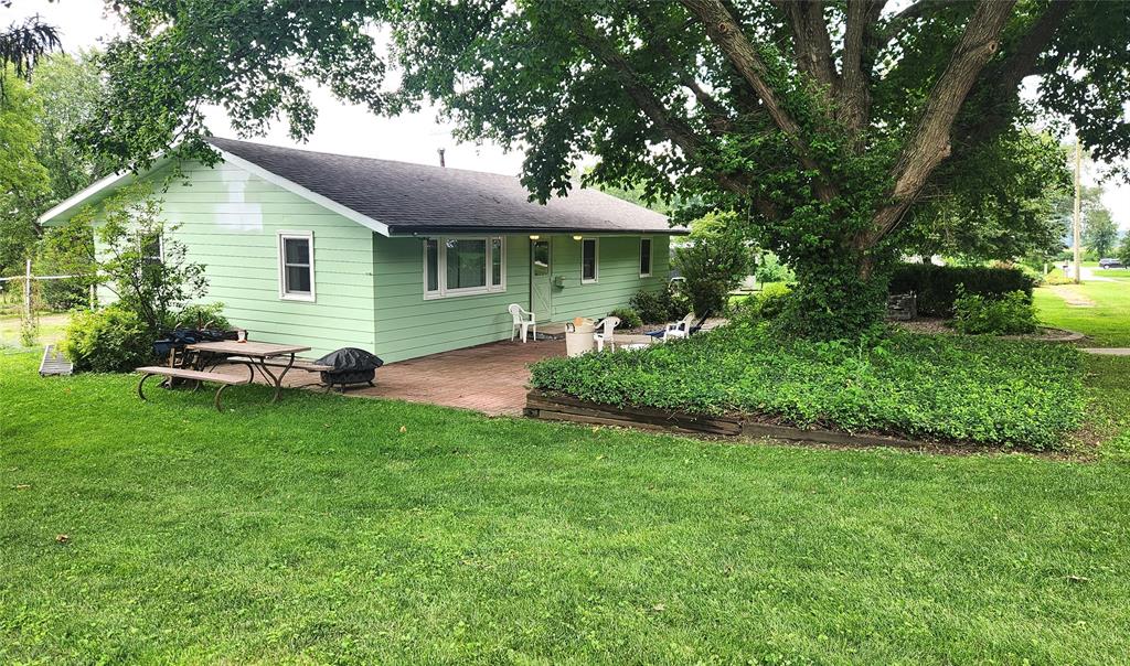 202 E Main St 34 A light green house with a dark roof sits next to a large tree and garden. There is a patio with white chairs and a picnic table on a grassy lawn, surrounded by lush greenery and leafy trees.