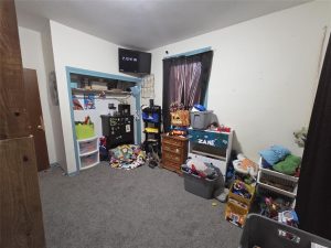 A cluttered child's bedroom with toys, bins, and storage shelves filling one side. A small TV is mounted high on the wall, and a window with dark curtains is above a dresser labeled "Zane." The carpet is gray.