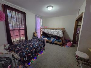 A cozy bedroom with a single bed on the left, a bunk bed on the right, stuffed animals, colorful bedding, string lights on the floor, and a window with curtains letting in natural light.