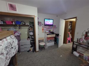 A child’s bedroom with a bunk bed, open closet, chest of drawers, TV on the wall displaying a cartoon, and various toys and clothes scattered around. The room has carpeted floors and colorful decorations.