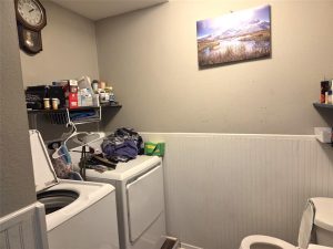 A laundry room with a washing machine, dryer, shelves holding cleaning supplies, clothes hanging to dry, a wall clock, a landscape picture on the wall, and part of a toilet visible.