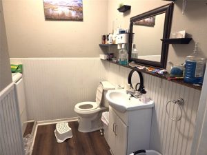 A small bathroom with a toilet, white sink vanity, shelves holding toiletries, a wall mirror, a step stool, a trash can, and a washing machine partially visible on the left. The floor is dark wood, and there is a landscape picture on the wall.