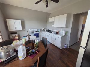 A kitchen with white cabinets and appliances, a wooden dining table cluttered with groceries and household items, and a ceiling fan overhead. The floor is dark wood, and a person is partially visible in the hallway.