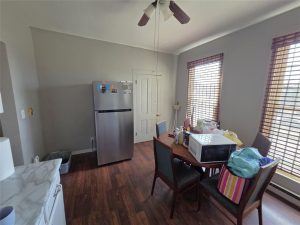 A small kitchen with a stainless steel refrigerator, a wooden table with four chairs, and various items including a microwave and bags on the table. Two large windows with blinds let in natural light.