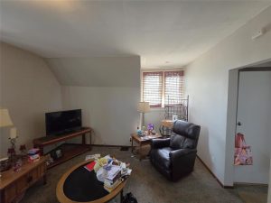 A cozy living room with a recliner, TV on a wooden stand, coffee table cluttered with papers, and two lamps. Light enters through two windows with blinds. A small pink bag hangs on a door handle.