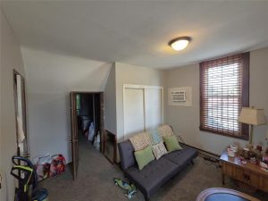 A small, carpeted room with a futon sofa, pillows, and a blanket. There is a wooden table with a lamp and assorted items, a window with blinds, and an air conditioner. An open door reveals another room.