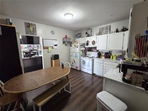 A cozy kitchen with white cabinets, a wooden table and bench, refrigerator, stove, and various kitchen items on the counters. Wall art, a bulletin board, and a bright window add character to the room.