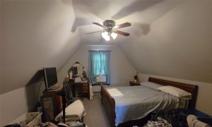 A cozy attic bedroom with sloped ceilings, a ceiling fan, a made bed, a TV on a dresser, a vanity with a mirror, and a window with blue curtains letting in daylight. Clothes and items are visible on the floor and furniture.