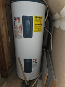 A tall, cylindrical white water heater with labels and a yellow EnergyGuide sticker stands in a narrow closet with exposed pipes and wires nearby. A wooden stick and two panels rest against the heater.