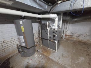 A basement with a water heater and a furnace unit next to each other; exposed pipes and ducts run along the ceiling and walls, which are made of painted bricks. The floor is bare concrete with some stains.