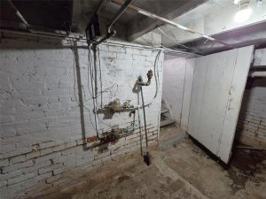 Dimly lit basement with exposed pipes and wiring on white brick walls, rough concrete floor, and an open wooden door leading to a small stairway. The area appears old and somewhat neglected.