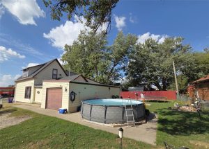 A round above-ground pool with a ladder sits in the backyard of a tan house with a garage. The yard has trees, a red fence, and scattered outdoor items, all under a bright blue sky with clouds.