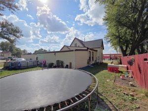 A backyard with a large trampoline, a beige house, a red fence, a small playhouse, trees, and a partly cloudy sky on a sunny day.