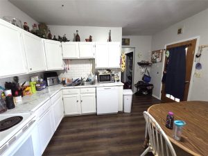 A kitchen with white cabinets, a white tile backsplash, and wood flooring. Various items are on the counters and above the cabinets. A small table with chairs and water bottles is in the foreground. A door and entryway are in the background.