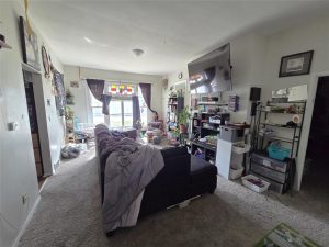 A cozy, lived-in living room with various plants, a black couch, bookshelves, a mounted TV, a large window with colorful stained glass, and shelves filled with books and storage bins. Sunlight brightens the space.