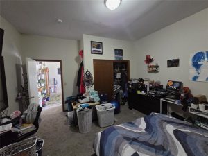 A cluttered bedroom with a bed in the foreground, two laundry baskets on the floor, clothes and items on dressers, an open closet, and a door leading to a brightly lit hallway. Various decorations and artwork are on the walls.