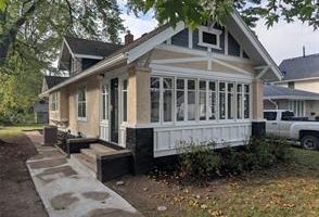 A beige and white craftsman-style house with a large enclosed front porch, black stone accents, and a small front garden, surrounded by trees and a neighboring house. A white pickup truck is parked nearby.