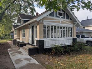 A beige and white craftsman-style house with a large enclosed front porch, black stone accents, and a small front garden, surrounded by trees and a neighboring house. A white pickup truck is parked nearby.