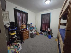 A cluttered bedroom with gray carpet, two windows with dark curtains, a wooden bunk bed, plastic storage bins, toys scattered on the floor, a small dresser, and a shelf filled with various items.