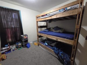 A small bedroom with gray carpet, a wooden triple bunk bed with blue bedding, and a window covered by dark curtains. Toys and storage bins are arranged against the walls.