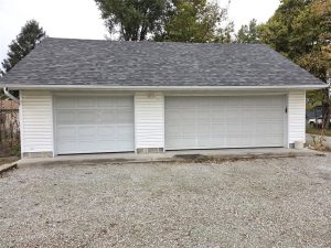 A detached, white, two-car garage with two closed light gray doors, a shingled roof, and a gravel driveway in front, surrounded by some trees and greenery.