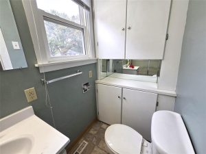Small bathroom with white cabinets, a sink, and a toilet. A window lets in natural light. There are mirrors above the countertop and a towel bar on the wall. The walls are painted light blue.