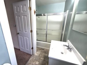 Small bathroom with a white vanity sink, a mirrored wall, a bathtub with frosted sliding glass doors, and a door leading to a room with carpeted flooring. The walls are light blue.