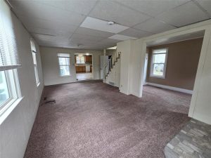Spacious living room with brown carpet, white walls, and multiple windows, leading to an open kitchen and a staircase. Separate room with large window is visible on the right side.