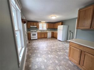 Spacious kitchen with wood cabinets, white appliances including a stove, dishwasher, and refrigerator, large windows on the left, and light-colored tile flooring. Walls are painted light blue-gray.