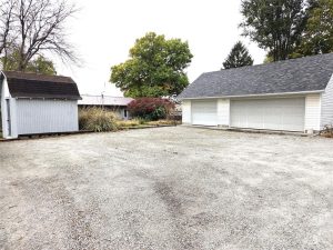 A gravel driveway leads to a large white garage with three closed doors on the right and a small gray shed on the left, surrounded by trees and shrubs under an overcast sky.
